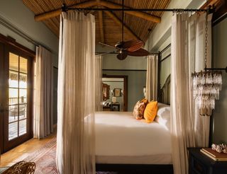 Elegant bedroom with a four-poster bed draped with sheer white curtains, accented by decorative pillows and a vintage chandelier, featuring a bamboo ceiling and a view to a balcony through open wooden doors.