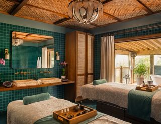 Spa treatment room with two massage tables, a rustic stone countertop with sinks, and a view of a balcony, featuring a woven wood ceiling, teal tile walls, and a large mirror.