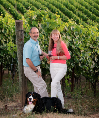 Couple holding wine with their dog and a winery in background