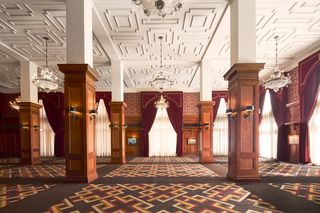 LAAC Centennial Ballroom event space with ornate, classical interior. Featuring white pillars, crystal chandeliers, dark wood paneling, patterned carpet, and red velvet drapes.