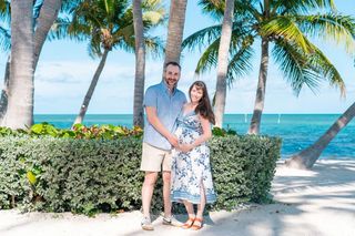 A pregnant woman in a floral dress and a man in a blue shirt and khaki shorts pose together, holding the woman's pregnant belly in front of a tropical beach background with palm trees, a green hedge, and ocean visible.