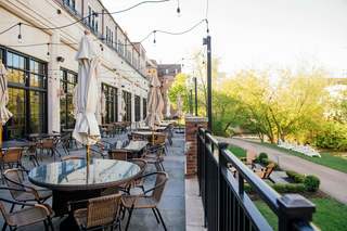 Outdoor restaurant patio with tables, chairs, and closed umbrellas, string lights overhead, with a black metal fence and view of trees and a pathway with chairs.