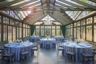 Interior shot of an ornate dining room with a glass ceiling and surrounding windows. Round tables covered in light blue tablecloths are set for a formal event with silverware, glassware, and floral centerpieces, with matching chairs. A chandelier hangs above the center tables, and a fountain can be seen through the windows at the back.