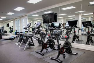 Gym interior with multiple exercise machines including stationary bikes, elliptical machines, and rowing machines, mirrored wall, and a dark grey rubber floor. Two Peloton stationary bikes are in the foreground.