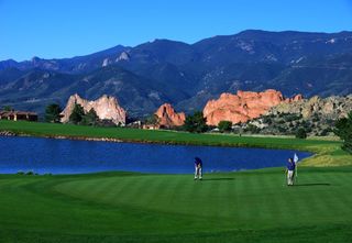 Two men playing a game of golf outside on a putting green with a body of water and mountains in the background.