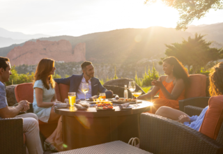Group of people seated outside around a table full of bites and drinks with the sun shining and mountains in the background.