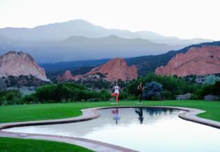 Two women holding a standing yoga pose outside in front of a tranquil pool with mountain views.