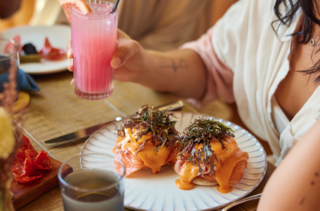 A woman dining at The Aster, holding a pink drink with a plate of eggs benedict in front of her and a charcuterie board on the side.
