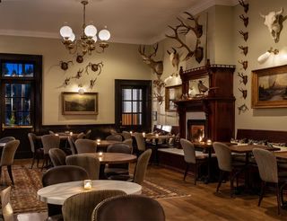 Interior of a dimly lit restaurant or bar, featuring round wooden tables with lit candles, upholstered chairs, a fireplace with a decorative mantelpiece adorned with taxidermy, paintings, and antlers on the walls, and a chandelier hanging from the ceiling.