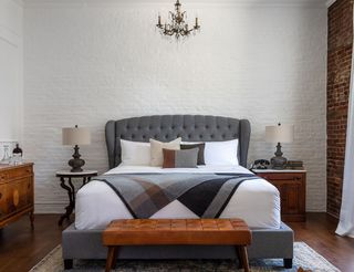 A stylish bedroom with a large, tufted grey headboard, white bedding, and patterned pillows. A grey and brown plaid blanket is draped over the bed. Two ornate lamps sit on bedside tables, and a brown leather bench is at the foot of the bed. A chandelier hangs above the bed. The backdrop is a white-painted brick wall with a exposed brick accent.