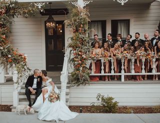 A bride and groom sit on porch steps with a small dog, while a large wedding party stands behind them holding bouquets on the porch decorated with flowers.