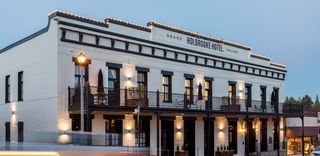 Exterior view of the Holbrooke Hotel in Grass Valley, California, a three-story, white brick building with black trim, balconies, and windows, illuminated by soft lighting at dusk. The hotel's name is displayed across the top of the building, and small lights line the roof.