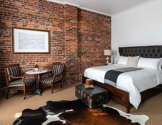 A hotel room with exposed brick walls, a dark wood framed bed with a tufted headboard, and a cowhide rug. Two striped armchairs and a small table with drinks sit in front of the brick wall, along with a framed blueprint. A vintage trunk sits at the foot of the bed.