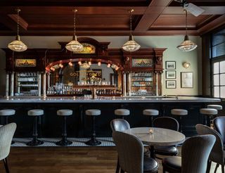 Interior view of a vintage bar, the "Golden Gate Saloon," featuring a long marble-topped bar with ornate dark wood detailing, pendant lights, and bar stools, with a few small round tables with chairs and a stained-glass and mirror back bar.