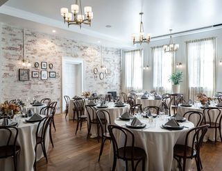 Elegant dining room set for a formal event with round tables draped in ivory linens, dark wooden chairs, and place settings featuring black and white striped napkins and floral centerpieces; exposed brick wall and multiple chandeliers illuminate the room.