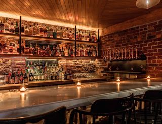 A dimly lit bar with shelves of liquor bottles, a marble counter, tap handles on the brick wall, and small candles placed on the countertop.
