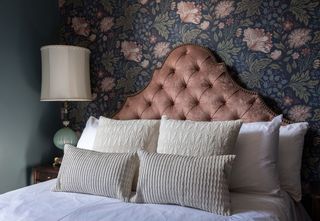 Bedroom interior with a tufted pink headboard, patterned floral wallpaper, and a bed with decorative pillows and a bedside lamp.