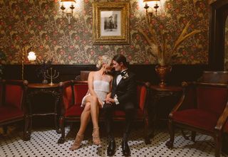 Newly married couple, a woman in a white fringe dress and a woman in a black tuxedo, sit closely on a red velvet couch, holding hands, in a vintage-style room with patterned wallpaper, ornate furniture, and decorative lighting.