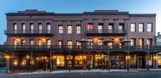 Three-story brick building with illuminated windows, balconies with ornate railings, and awnings over ground-level storefronts, lit by lampposts and overhead lights, under a twilight sky.
