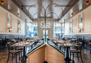 Interior view of a Lola with tables set for dining; a central teal-colored booth flanked by tables and chairs, under a dramatic brass light fixture and high ceilings, with an open doorway at the end of the room.