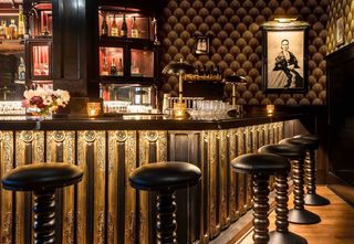 Interior of a dimly lit, art deco-style bar with a black, ornate bar counter featuring gold accents and black leather stools in the foreground, with liquor displays and an elaborate patterned wallpaper in the background.
