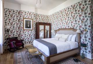 Bedroom with floral wallpaper, a large bed with a light-colored wood frame and neutral-colored bedding, a wooden armoire with mirrors, a burgundy rocking chair, two brass stools, and framed artwork.