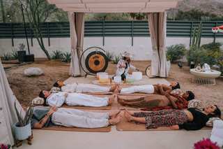 Seven people lie on yoga mats arranged in a circle, under a white canopy structure in an outdoor setting. A person dressed in a white shirt and hat, kneels near large crystal bowls and a large gold gong.
