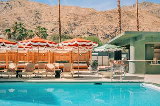 A luxurious outdoor pool area with clear blue water and lounge chairs beneath orange and white fringed umbrellas. In the background, there is a green bar with bar stools and a mountain range. There are lounge chairs with orange and white patterned pillows, as well as a row of wooden cabanas with seating. The scene is bright and sunny.