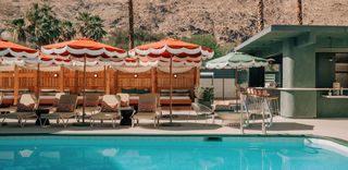 A luxurious outdoor pool area with clear blue water and lounge chairs beneath orange and white fringed umbrellas. In the background, there is a green bar with bar stools and a mountain range. There are lounge chairs with orange and white patterned pillows, as well as a row of wooden cabanas with seating. The scene is bright and sunny.