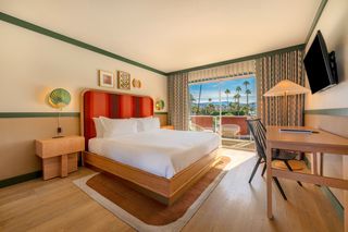 Interior view of a modern hotel room. A large bed with a red headboard and crisp white bedding dominates the center. A wooden nightstand with a green fan-shaped lamp sits on the left, next to the bed. A window opens to a view of palm trees, mountains, and blue sky. To the right is a small wooden desk, a black chair, and a wall-mounted television. The room has light wood floors and green trim on the walls and ceiling. The style is modern and minimalist.
