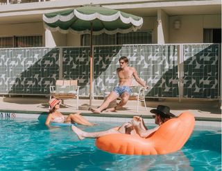 Three people enjoying the water and sun at a retro-style pool. A man shirtless, in blue patterned swim trunks, sits on a chair near the pool, while a woman wearing a sun hat and swimsuit floats on an inner tube. Another man, wearing a cowboy hat, is floating on an orange inflatable raft in the pool, holding a drink with a straw.