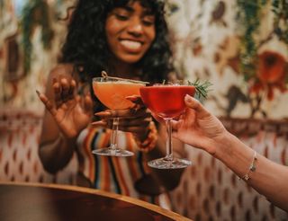 Two women in a restaurant clink cocktails; one woman holds a yellow drink and the other holds a red drink with rosemary garnish. The woman holding the yellow drink is smiling, wearing a striped dress; the other woman is wearing a light floral print top. The background is floral wallpaper and a wooden table.