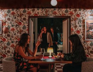 A dimly lit interior scene of High Moon restaurant with floral wallpaper. Two women sit at a wooden table facing each other, with drinks and snacks in front of them. A small lamp sits on the table between them, illuminating the scene. In the background, through an open doorway, a smiling woman and a man are seen entering the room. The wallpaper is a cream color with a floral design in shades of red, blue, green, and brown. Artwork, framed landscape scenes, hangs on the wall. The two women are seated in upholstered chairs.