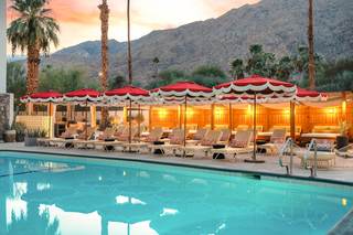 Royal Sun Palm Springs pool scene at sunset. The turquoise pool with a ladder is in the foreground, with rows of beige lounge chairs, each with a patterned pillow, and red and white fringe umbrellas lining its edge. Behind the lounge chairs is a wood-framed cabana with a canopy and warm lights. Tall palm trees are on the left, and a mountain range is in the background, with a colorful sky.