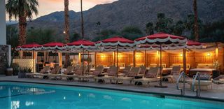 Royal Sun Palm Springs pool scene at sunset. The turquoise pool with a ladder is in the foreground, with rows of beige lounge chairs, each with a patterned pillow, and red and white fringe umbrellas lining its edge. Behind the lounge chairs is a wood-framed cabana with a canopy and warm lights. Tall palm trees are on the left, and a mountain range is in the background, with a colorful sky.