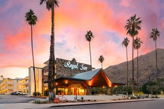 An exterior shot of the "Royal Sun" at sunset, featuring the neon sign "Royal Sun", with palm trees, a vibrant pink and orange sky, and a mountain in the background. The architecture includes a stone facade, a covered entrance, and a parking lot with a few parked cars. A decorative orange bicycle is in the foreground.