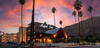 An exterior shot of the "Royal Sun" at sunset, featuring the neon sign "Royal Sun", with palm trees, a vibrant pink and orange sky, and a mountain in the background. The architecture includes a stone facade, a covered entrance, and a parking lot with a few parked cars. A decorative orange bicycle is in the foreground.