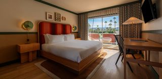 Interior view of a modern hotel room. A large bed with a red headboard and crisp white bedding dominates the center. A wooden nightstand with a green fan-shaped lamp sits on the left, next to the bed. A window opens to a view of palm trees, mountains, and blue sky. To the right is a small wooden desk, a black chair, and a wall-mounted television. The room has light wood floors and green trim on the walls and ceiling. The style is modern and minimalist.