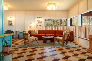 An interior shot of a brightly lit lobby lounge area with a terracotta-colored sectional sofa, two wooden chairs, and a round coffee table with a chessboard. There is a large window with a view of palm trees and cacti outside, draped by light brown and cream-colored curtains. The floor is covered in a geometric pattern of brown, beige, and dark brown tiles.