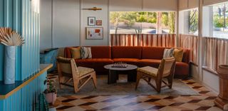 An interior shot of a brightly lit lobby lounge area with a terracotta-colored sectional sofa, two wooden chairs, and a round coffee table with a chessboard. There is a large window with a view of palm trees and cacti outside, draped by light brown and cream-colored curtains. The floor is covered in a geometric pattern of brown, beige, and dark brown tiles.