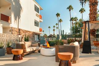 An outdoor lounge area with a mid-century modern aesthetic, featuring a fire pit, patio furniture, palm trees, and a two-story building in the background.