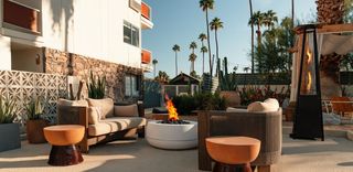 An outdoor lounge area with a mid-century modern aesthetic, featuring a fire pit, patio furniture, palm trees, and a two-story building in the background.