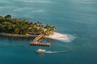 Aerial view of a tropical island with lush green vegetation, sandy beach, and luxury villas. A wooden pier extends into the turquoise water, and a blue and white boat leaves a white wake.