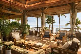 A luxurious tropical outdoor living space with wooden structure, thatched roof, and ocean views. Furnished with wicker furniture, large cushions, a wooden coffee table with an open book, and potted plants, creating a serene and inviting atmosphere.