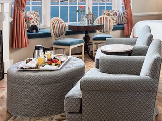 A luxurious hotel room interior with chairs, a round table, and an ottoman, all overlooking a seascape view through a window, with a tray of breakfast food and a bottle of wine on display.