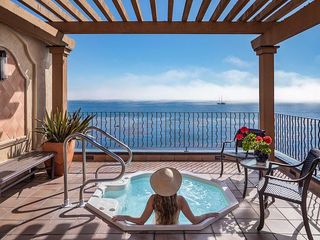 Woman in a hat relaxing in a rooftop hot tub overlooking the ocean and a distant sailboat, framed by a pergola and a wrought-iron railing.