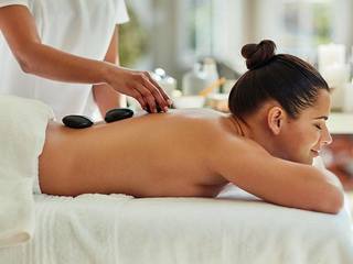Woman receiving a stone massage while lying face down on a massage table, with eyes closed in relaxation. A masseuse places hot stones on her back.