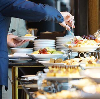 A person in a blue suit uses serving tongs to place food onto a white plate at a buffet table, featuring assorted fruits and pastries.