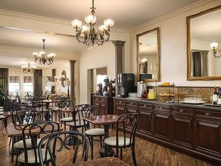 Interior of a hotel dining area with multiple small round tables, black bistro chairs, a coffee and drink station, gold-framed mirrors, and ornate chandeliers.