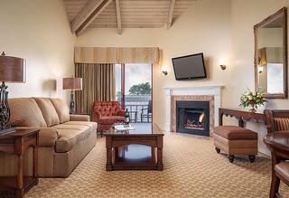 Elegant living room interior featuring a brown sofa, red patterned armchair, fireplace with fire, mounted TV, large mirror, and open doors leading to a balcony with an ocean view.
